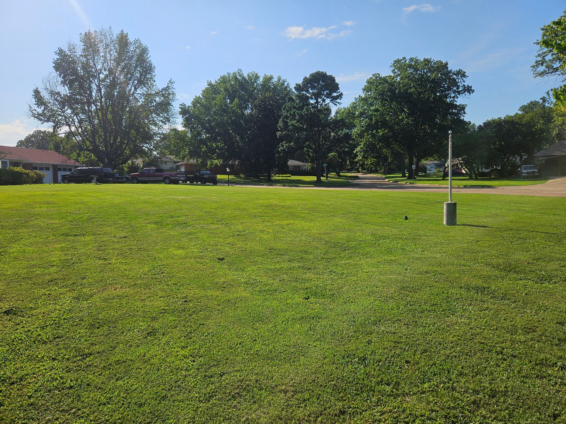 Sunny park with a wide grassy field, trees, a sidewalk, and a small utility box under a blue sky.
