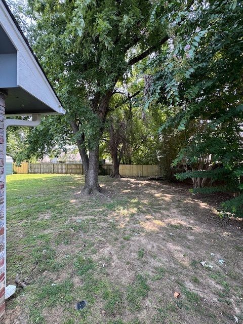 Shaded backyard with trees, patchy grass, and a wooden fence beside a house.