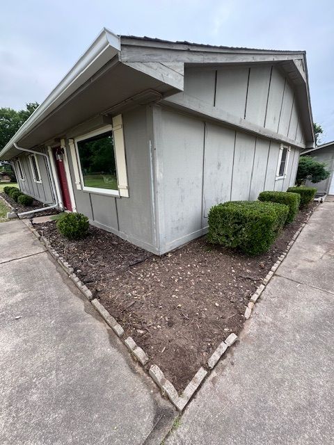 Corner view of a gray house exterior with white trim, small shrubs, and a concrete walkway