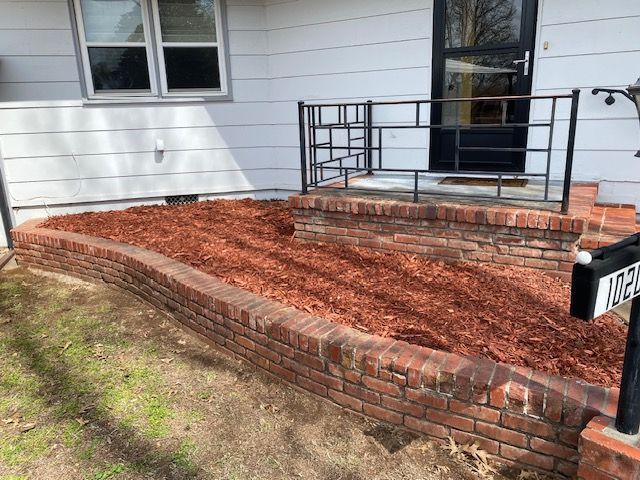 Brick planter bed with red mulch and black metal railing beside a house entrance