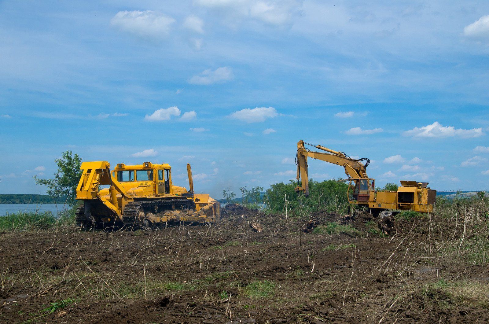 Two yellow construction vehicles clearing brush in a muddy field under a blue sky