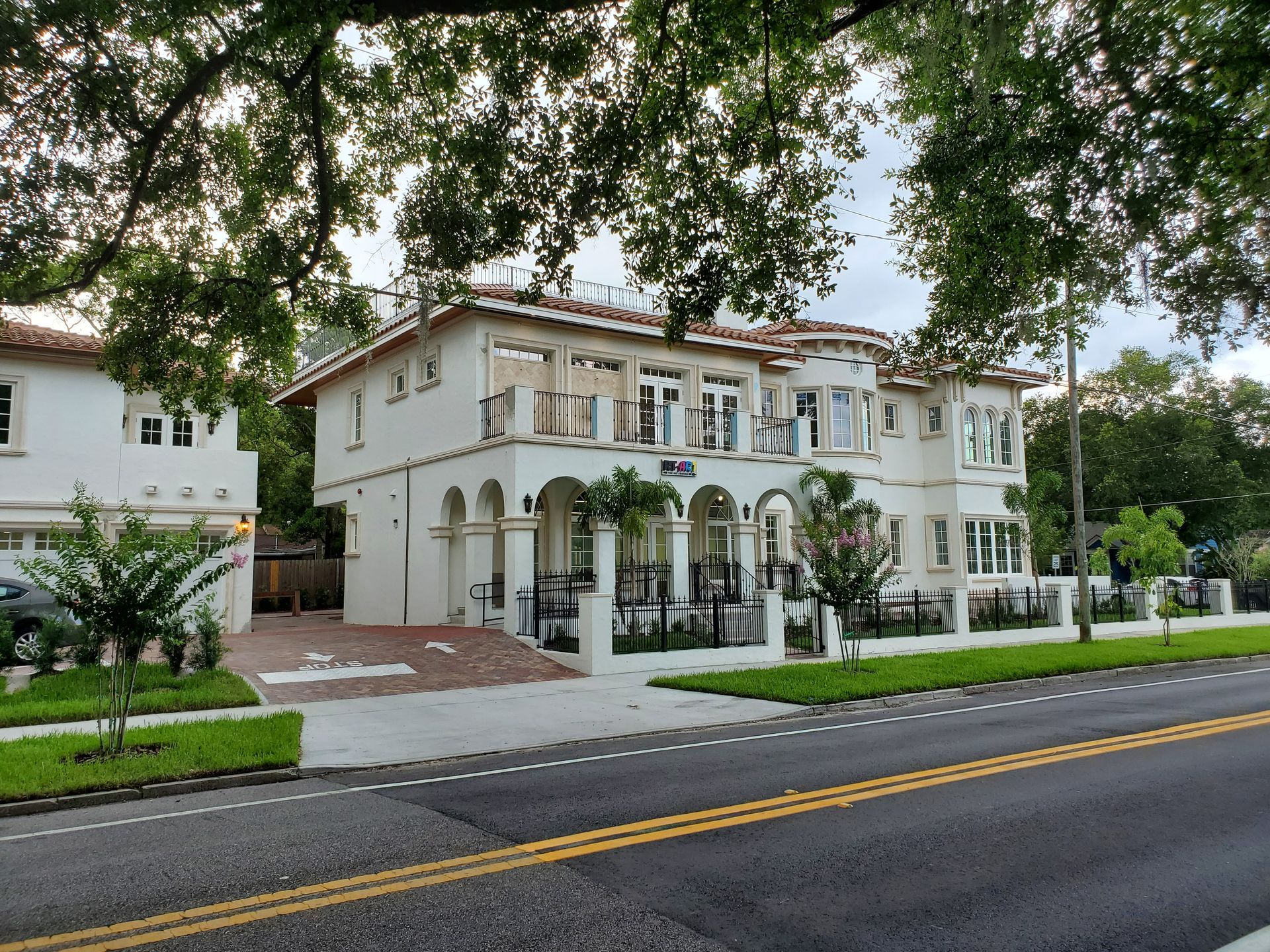 White colonial-style building with arches and balconies beside a road, framed by trees and greenery