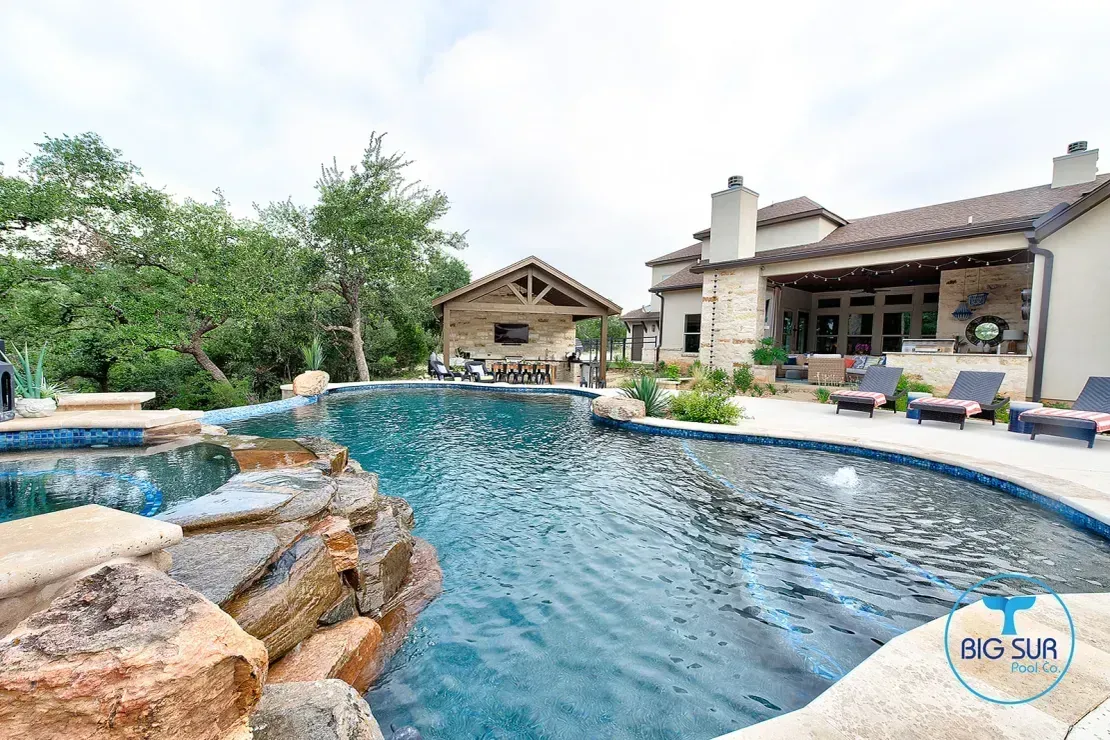 Backyard resort pool with rock waterfall and patio beside a stone house