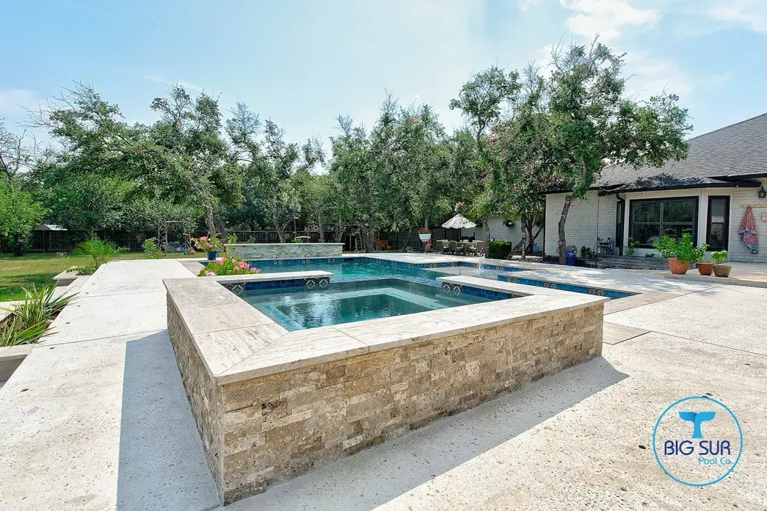 Backyard pool with stone raised edge, patio, and trees under a bright sky