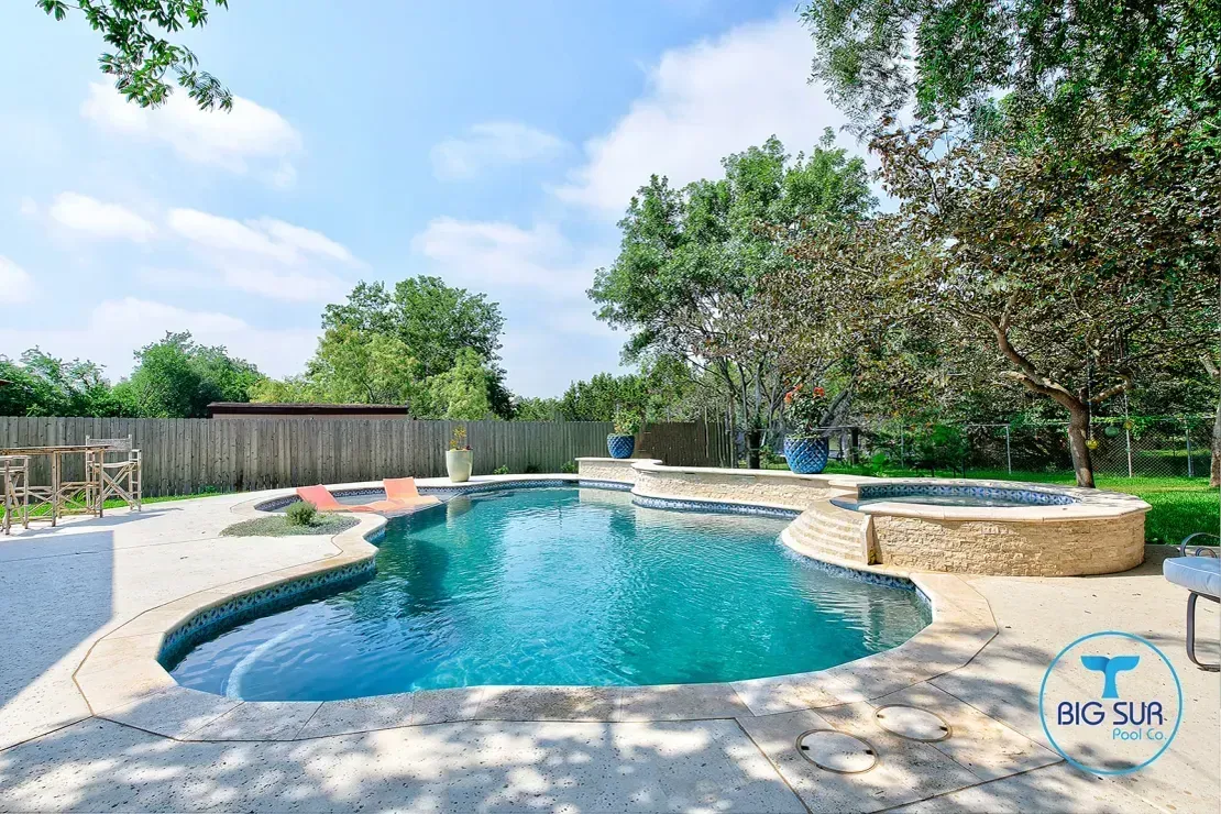 Backyard pool with curved blue water, stone patio, lounge chairs, and trees under a sunny sky