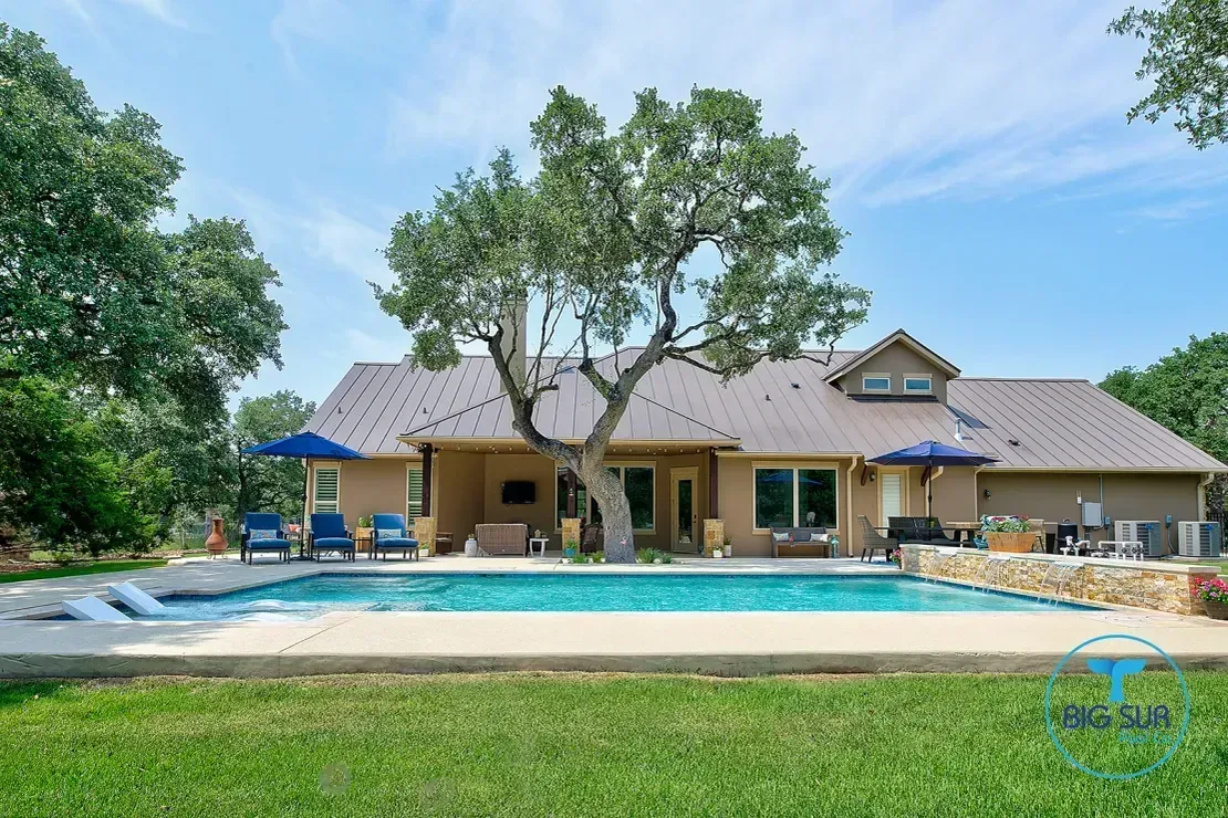 Suburban house with a large backyard pool, patio seating, and a tree in the front yard under a blue sky