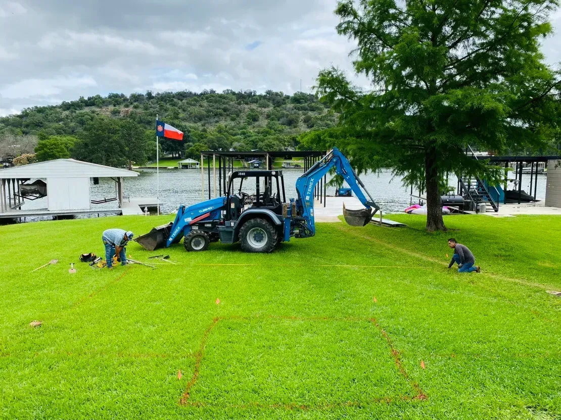 Blue tractor with backhoe on a grassy lakeside lawn, workers nearby and boats in the background