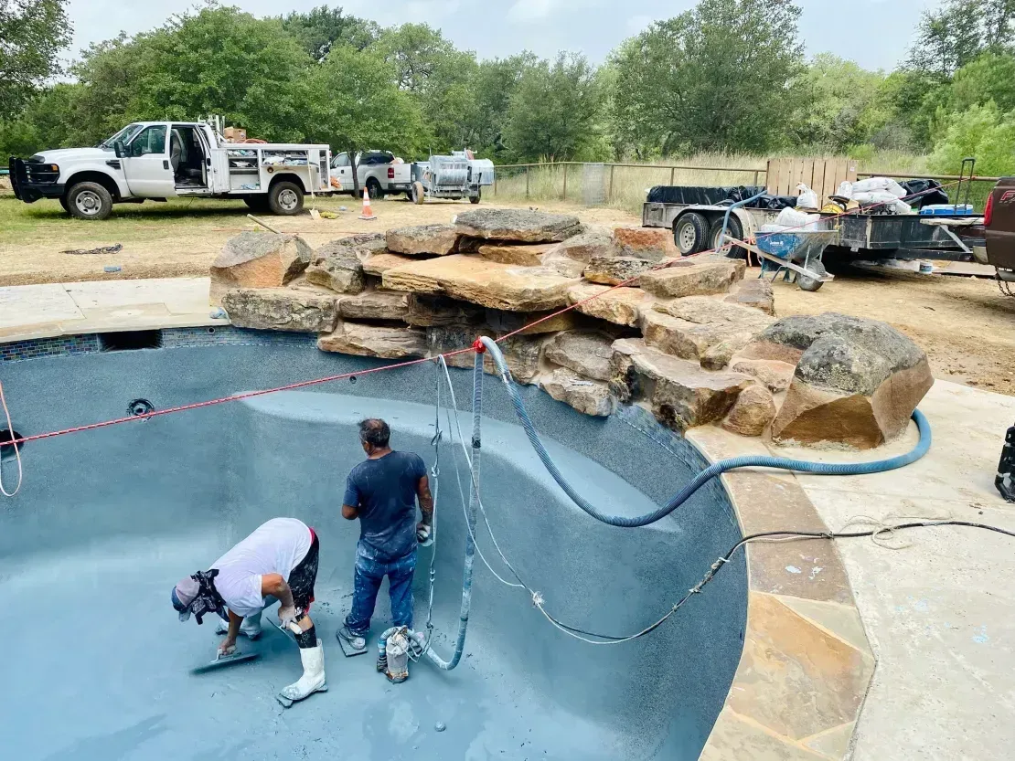 Workers cleaning an empty pool beside a rock waterfall and parked trucks outdoors