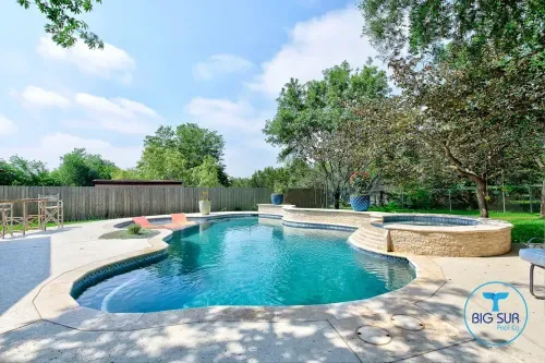 Backyard pool with blue water, stone patio, trees, and a hot tub under a bright sky