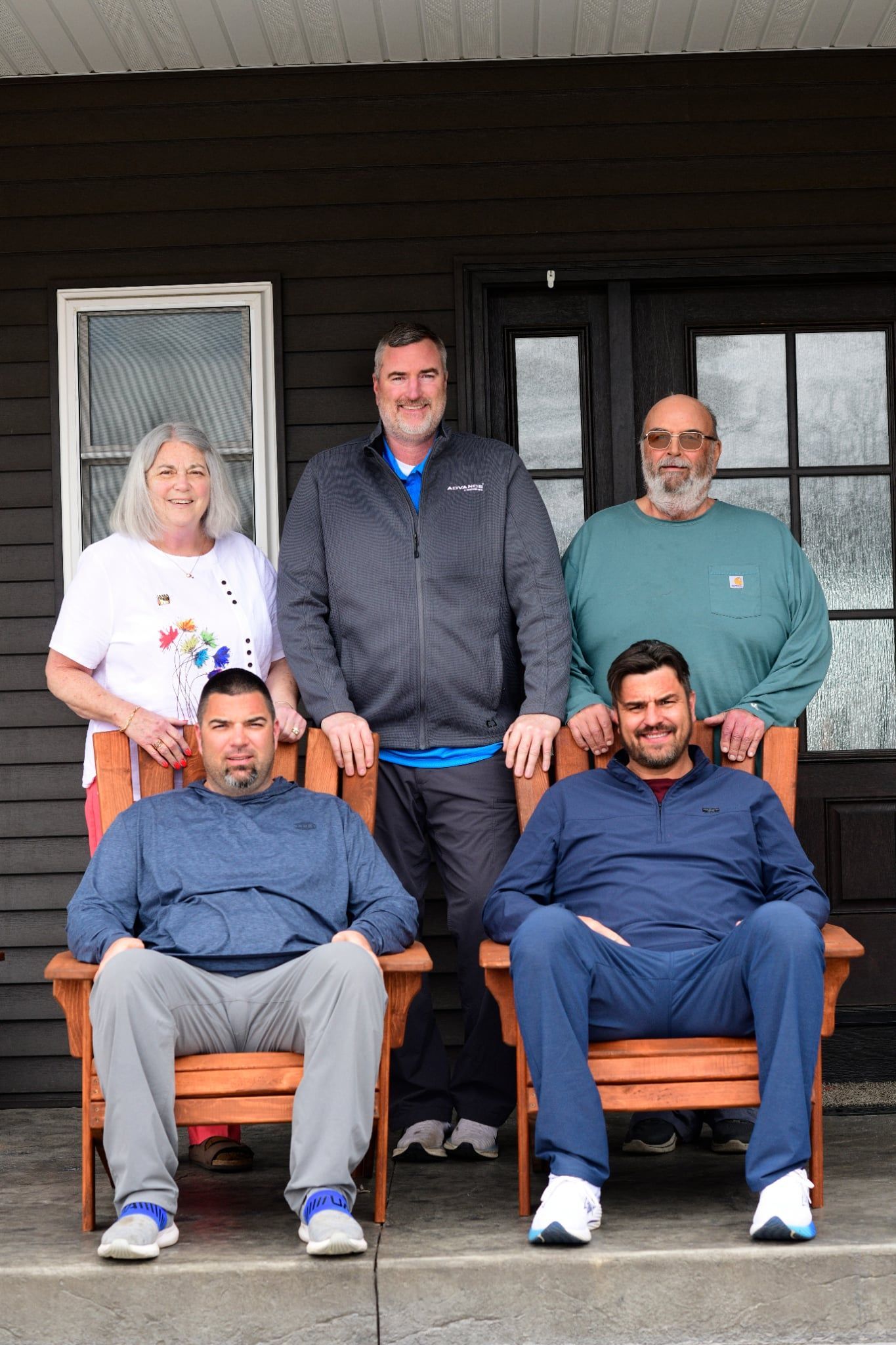 Five people posing on a porch; two seated in wooden chairs, three standing behind them.
