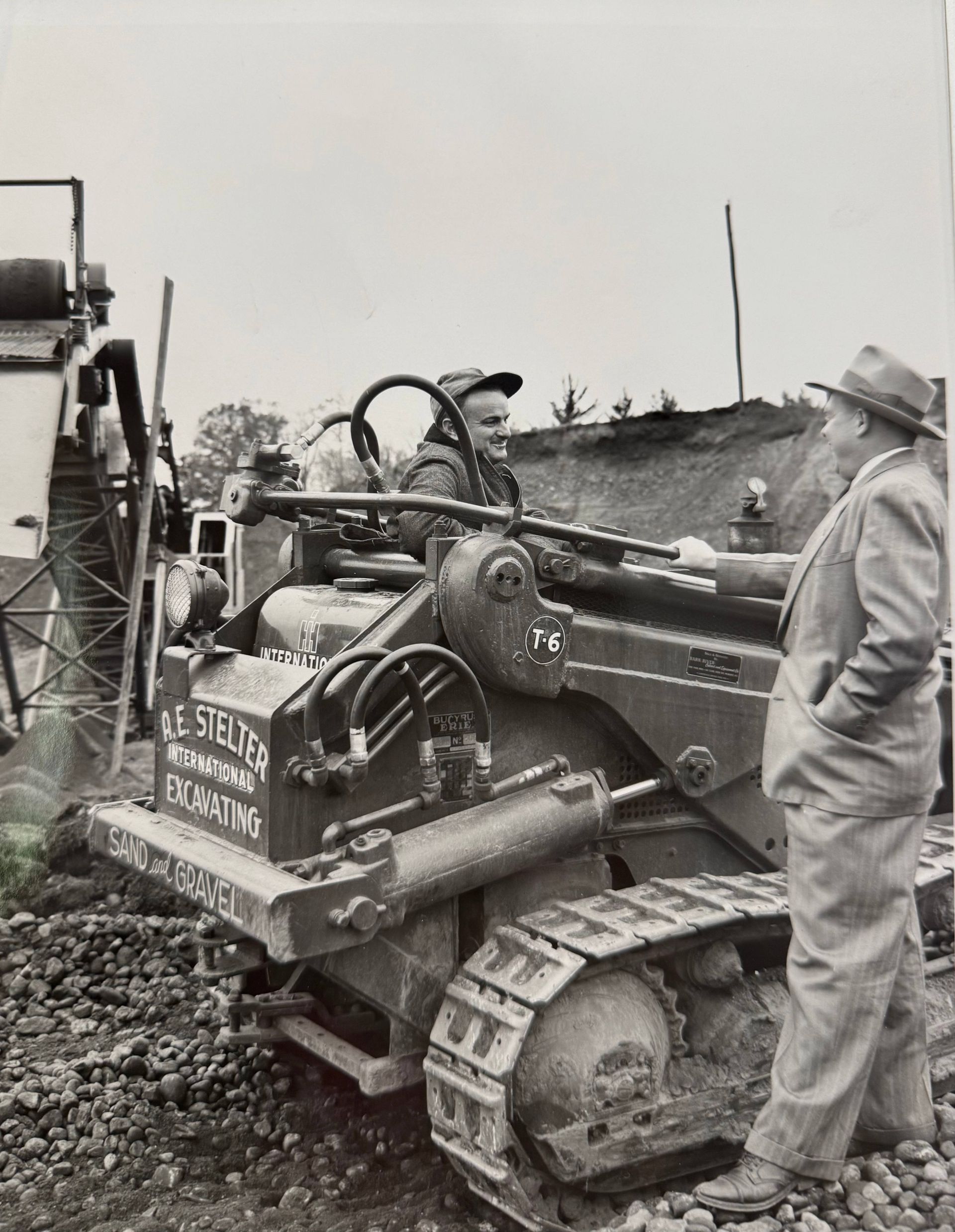 Man operating a small bulldozer on a construction site. Another man in a suit looks on.