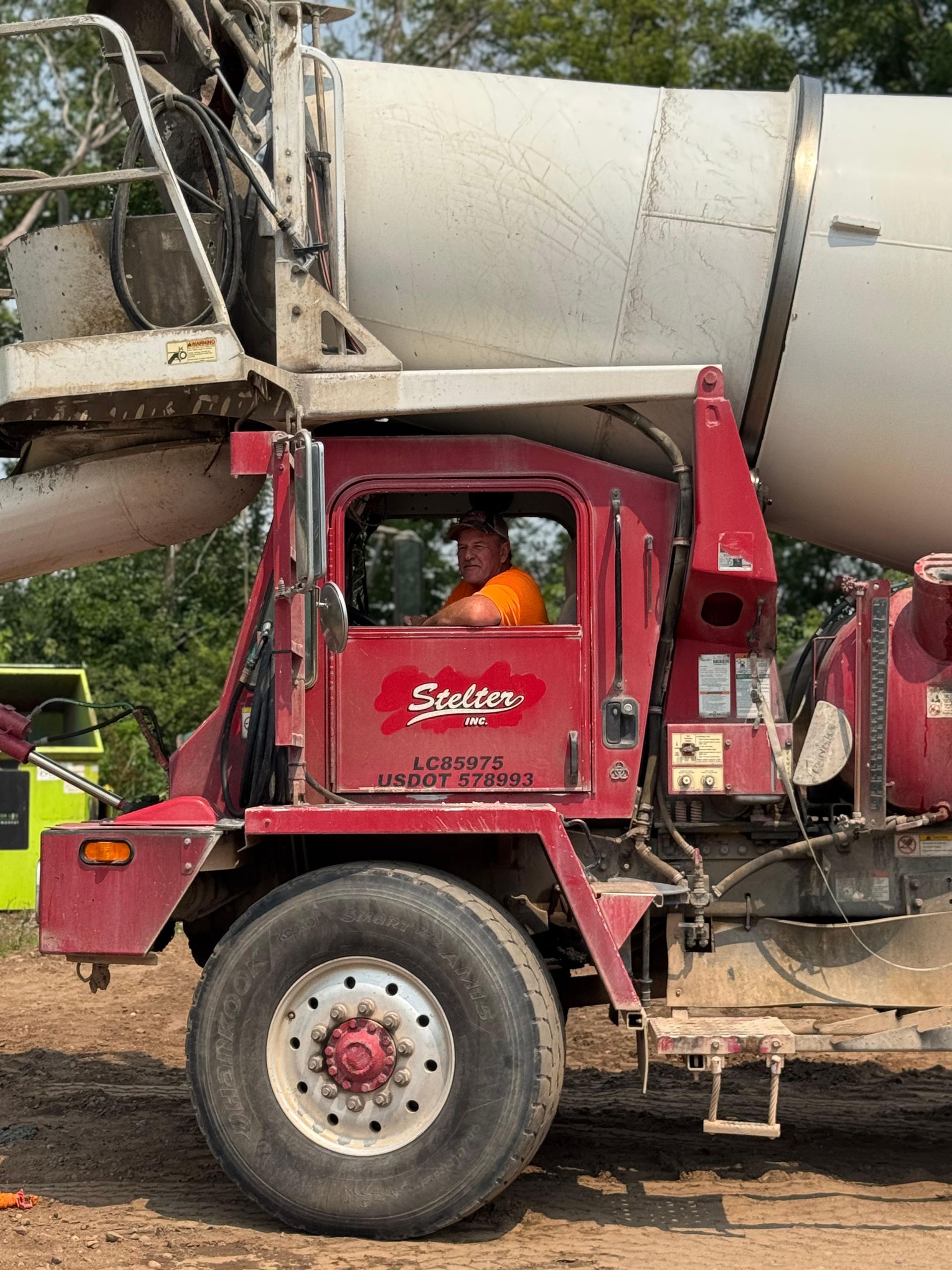 Cement truck with a driver in the cab, red truck, white drum, parked outside, green trees in the background.