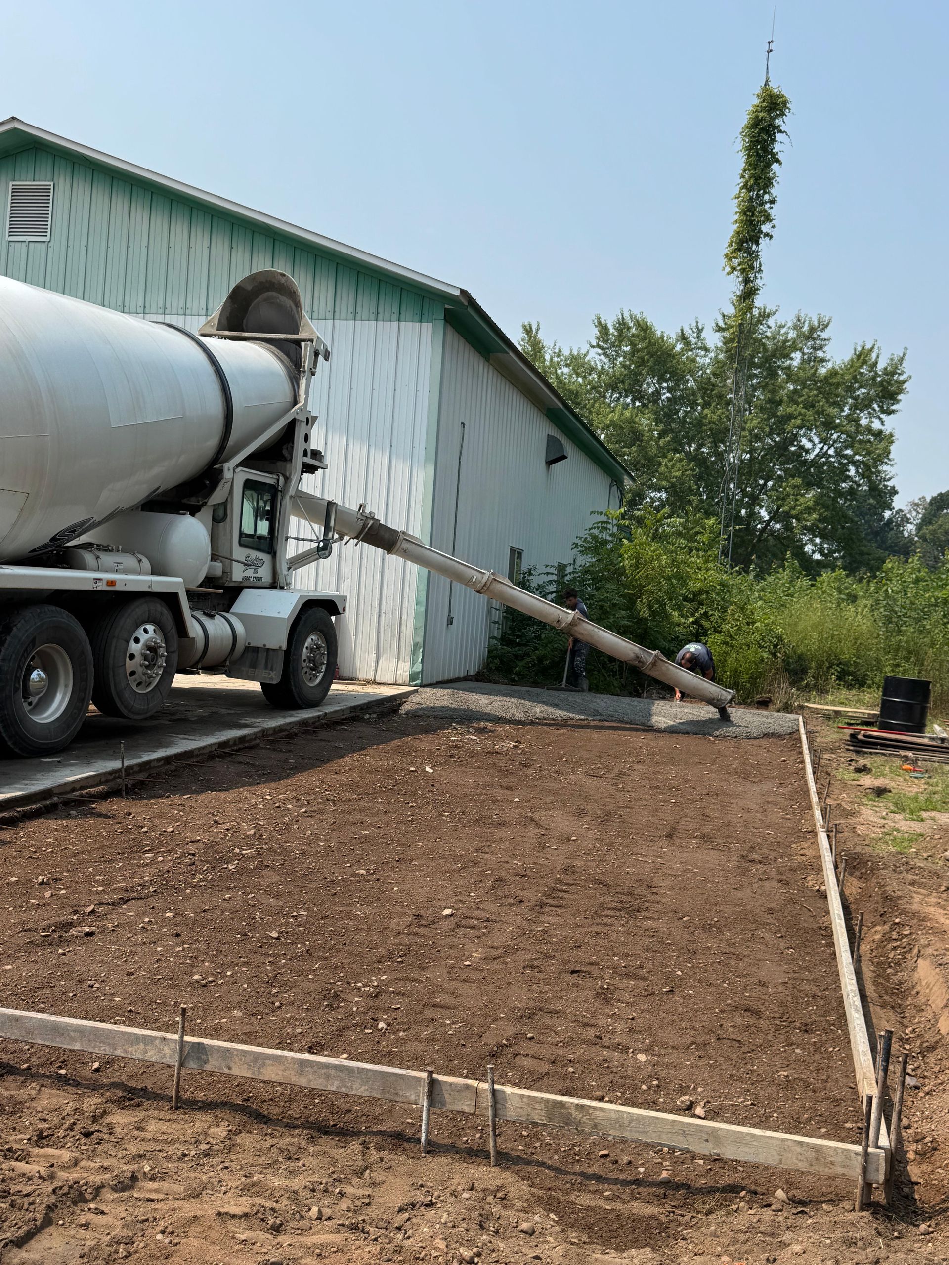 Concrete truck pouring cement into a framed area at a building's side on a sunny day.