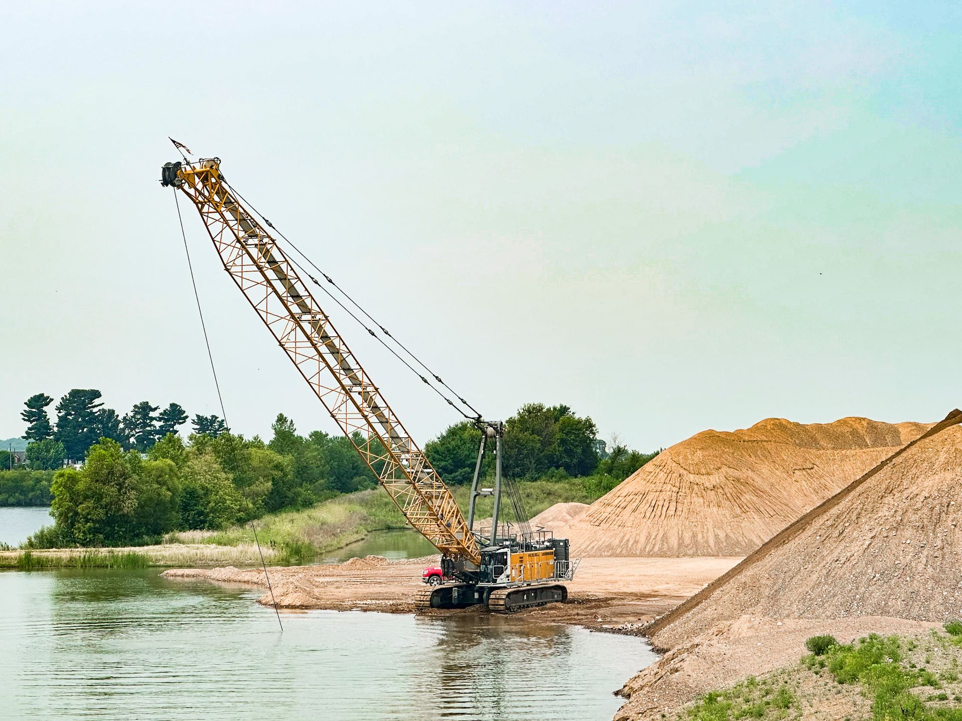 A large yellow crane scooping sand from a lake; piles of sand are visible on the shore.