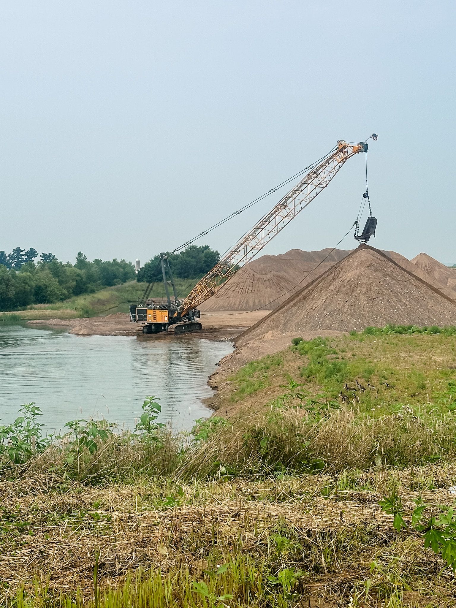 Crane scoops sediment from water, depositing it onto a pile on shore.