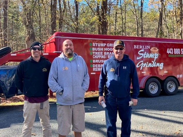 Three men are standing in front of a red trailer that says Berlin Stump Grinding			.