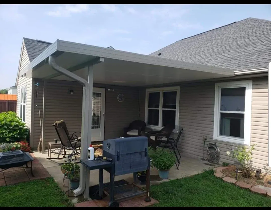 Covered backyard patio with grill, patio chairs, and house exterior in daylight