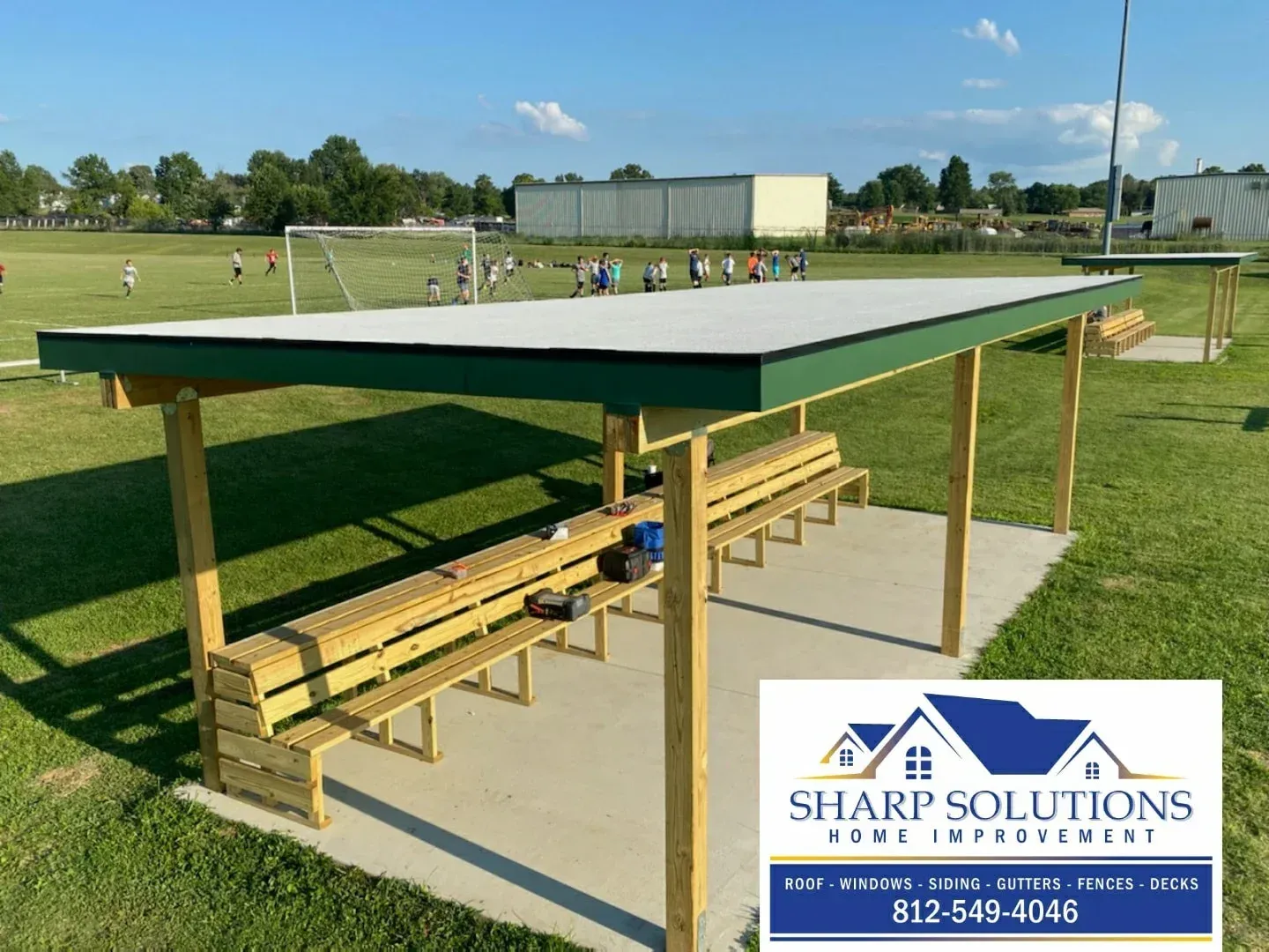 Covered outdoor picnic shelter with yellow benches on a sports field, with a Sharp Solutions logo in the corner