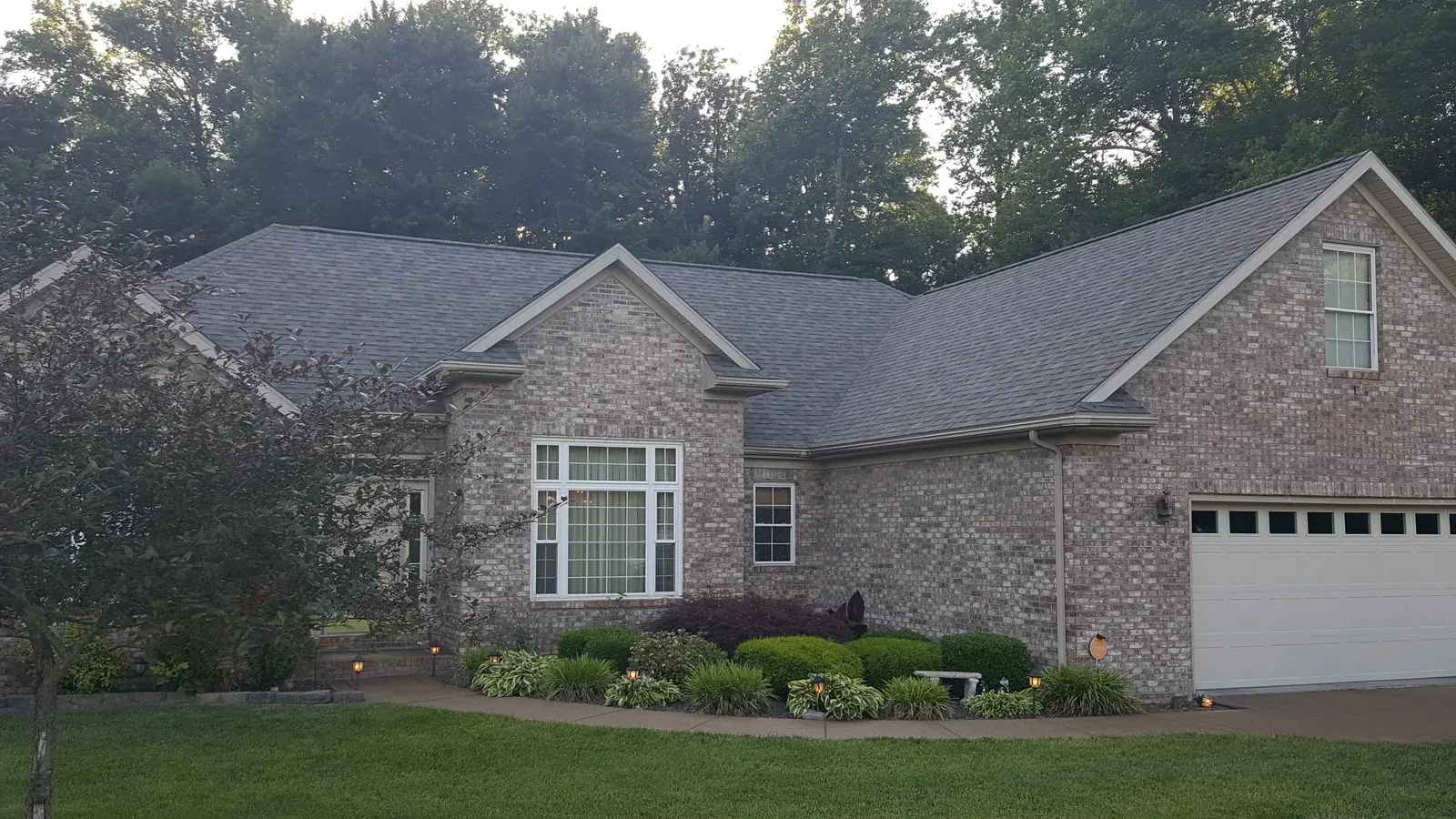 Brick suburban house with dark roof, white garage door, and landscaped front yard at dusk