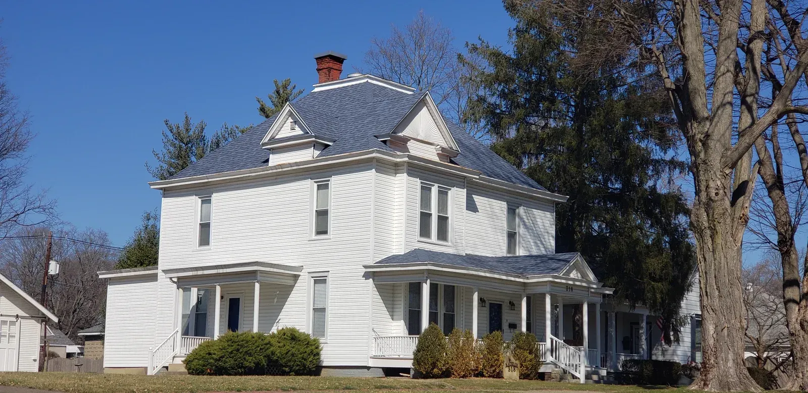White two-story house with a front porch, dark shutters, and trees in a sunny yard