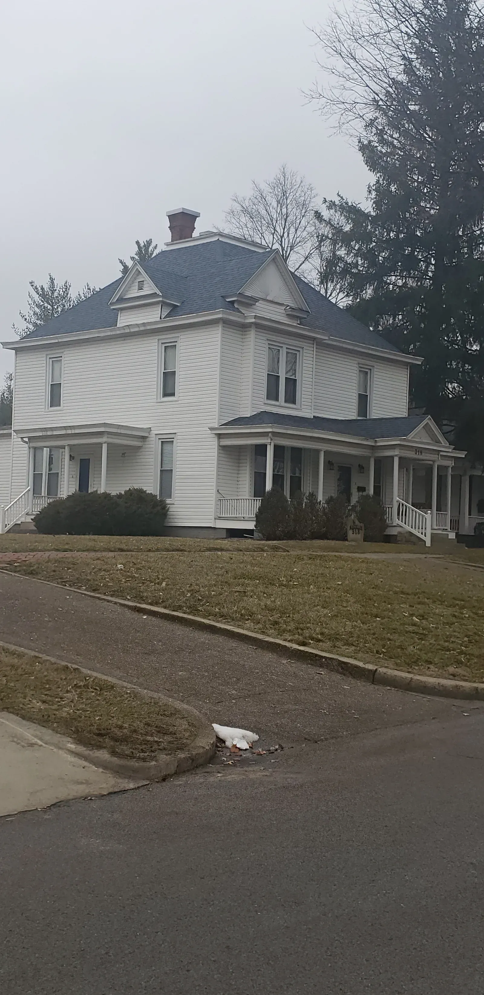 White Victorian-style house on a corner lot with a steep roof, porch, and bare trees on a gray day