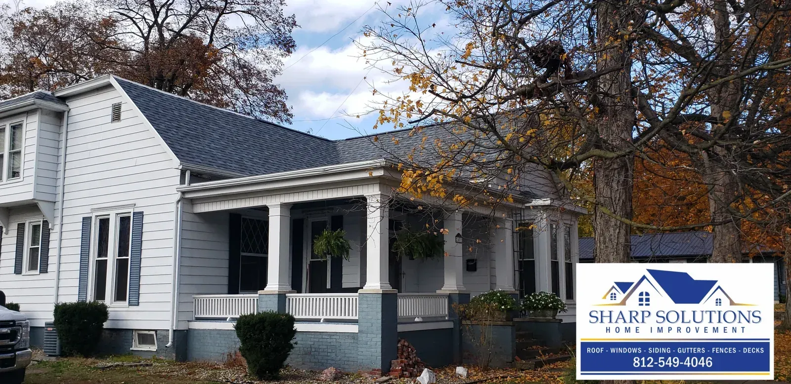 White house with dark roof and porch under autumn trees, with a small business sign in front
