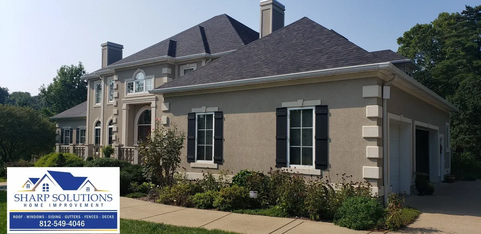 Large beige house with dark roof, stone trim, and green shutters, with a landscaping company sign in front