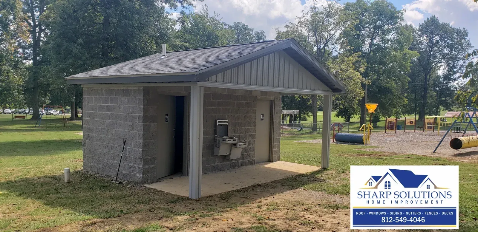Small gray stone pavilion with a gabled roof and open side, set in a grassy park near a fence and trees.