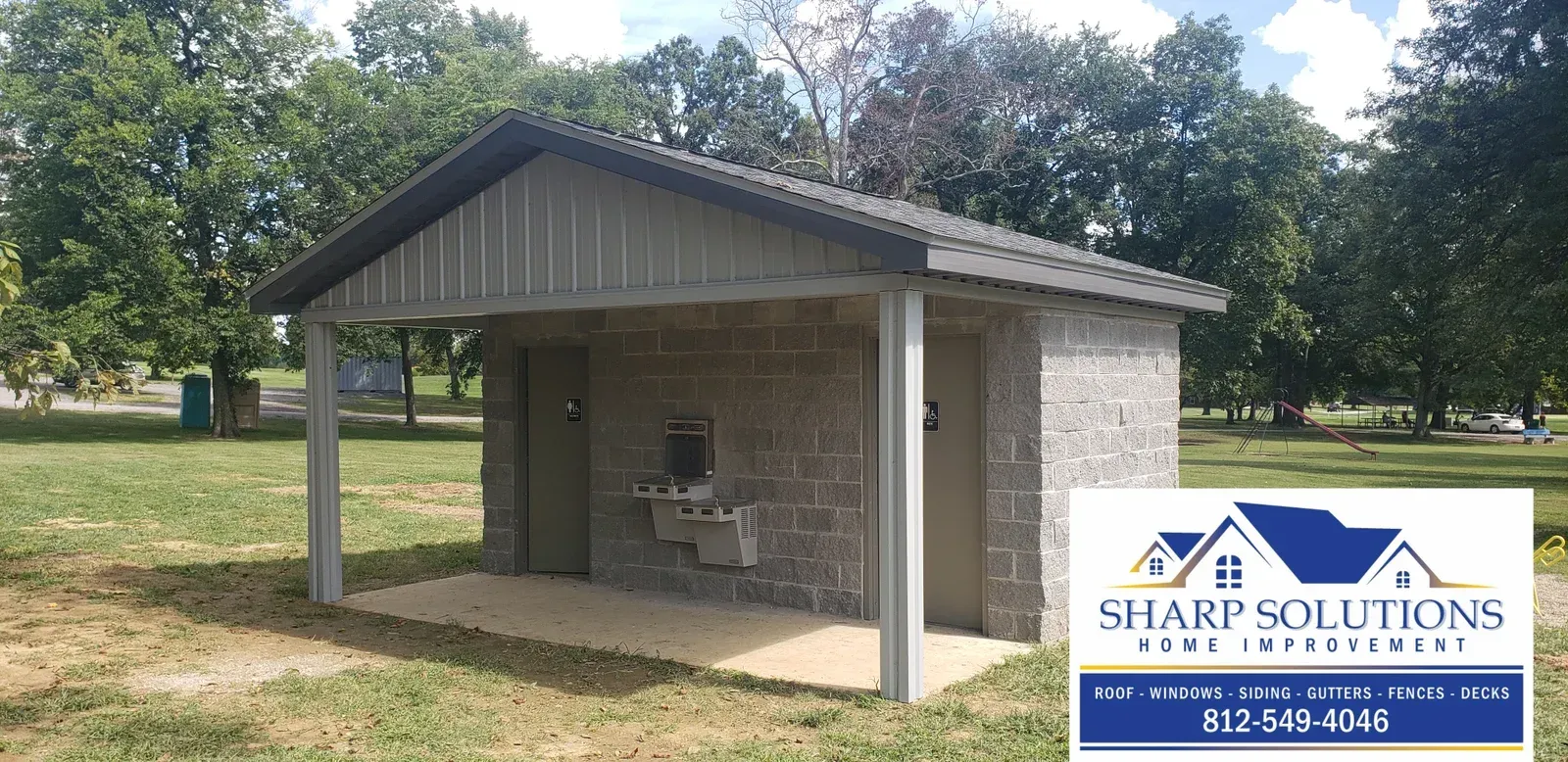 Small open pavilion with a metal roof and concrete posts in a grassy park, with a Sharp Solutions sign at right