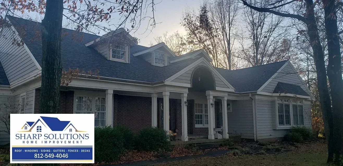A white house with a front porch and dark roof in a wooded yard at dusk, with a small logo in the corner