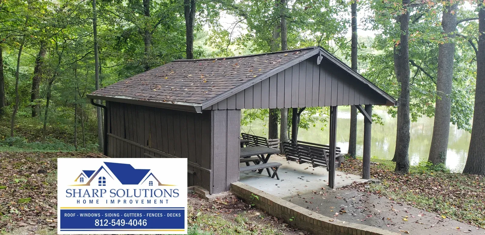 Small dark wooden pavilion with stone floor in a wooded park, next to a Sharp Solutions sign.