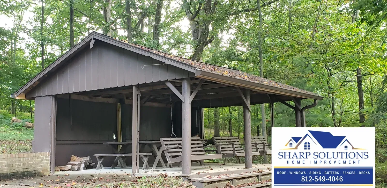 Gray open-sided pavilion in a wooded area, with stacked firewood and a “Sharp Solutions” sign in front