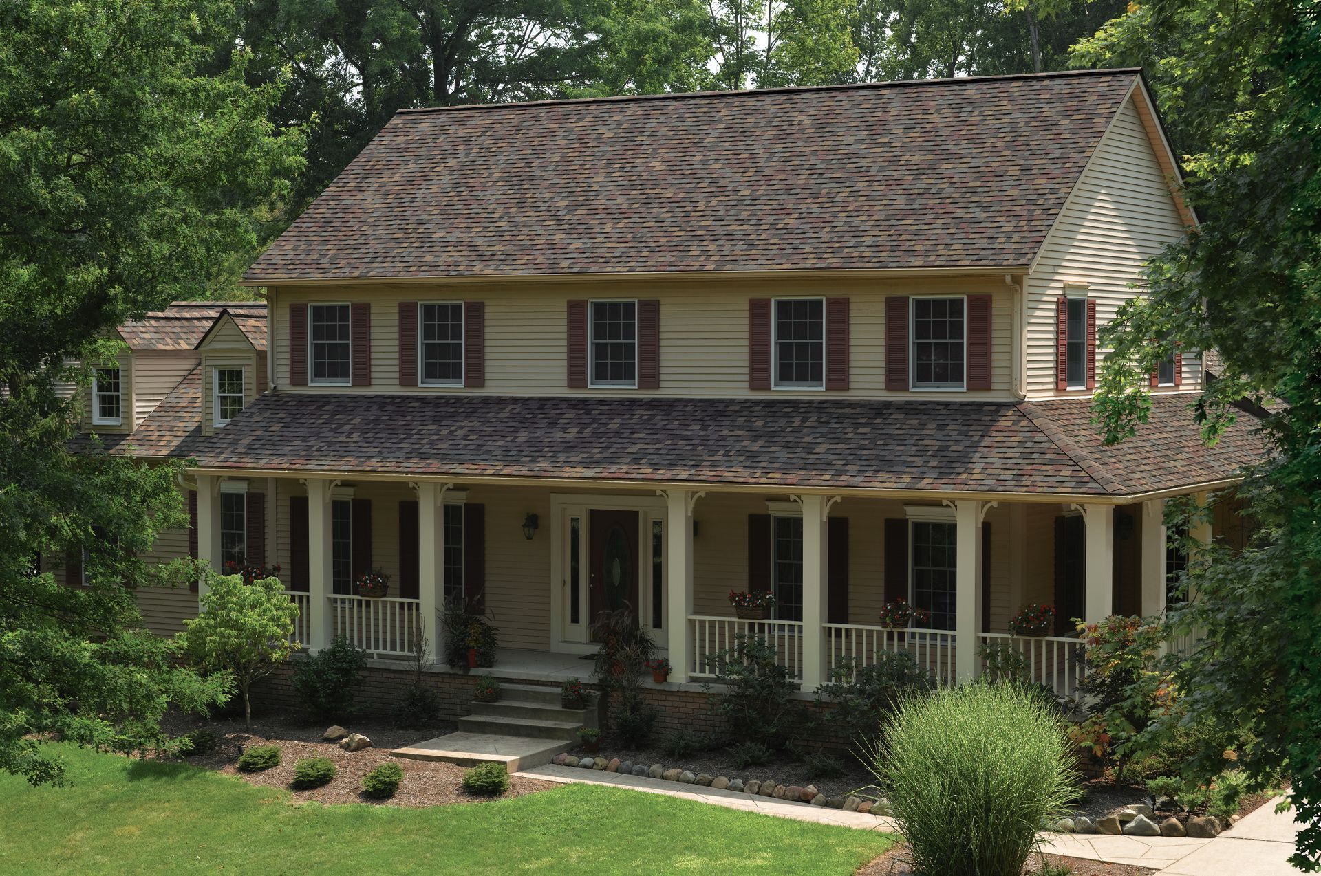 Two-story beige house with a covered porch, brown roof, and green trees and lawn