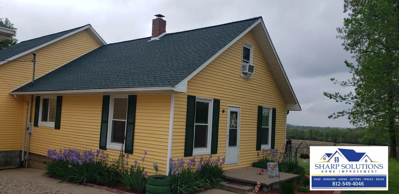 Yellow cottage with dark roof and white trim, next to a larger house under an overcast sky.