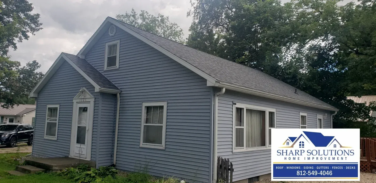 Blue-gray house with white trim and a steep roof, surrounded by trees and a driveway