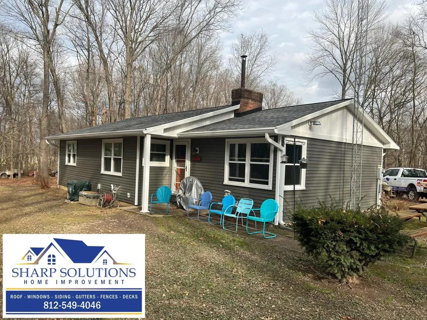 Small gray house with white trim and blue chairs on a porch in a wooded yard, with a Sharp Solutions sign.