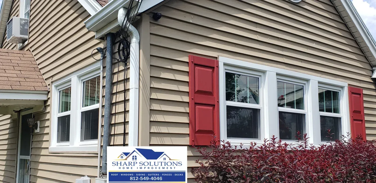 Suburban house exterior with beige siding, red shutters, and a realty sign in front