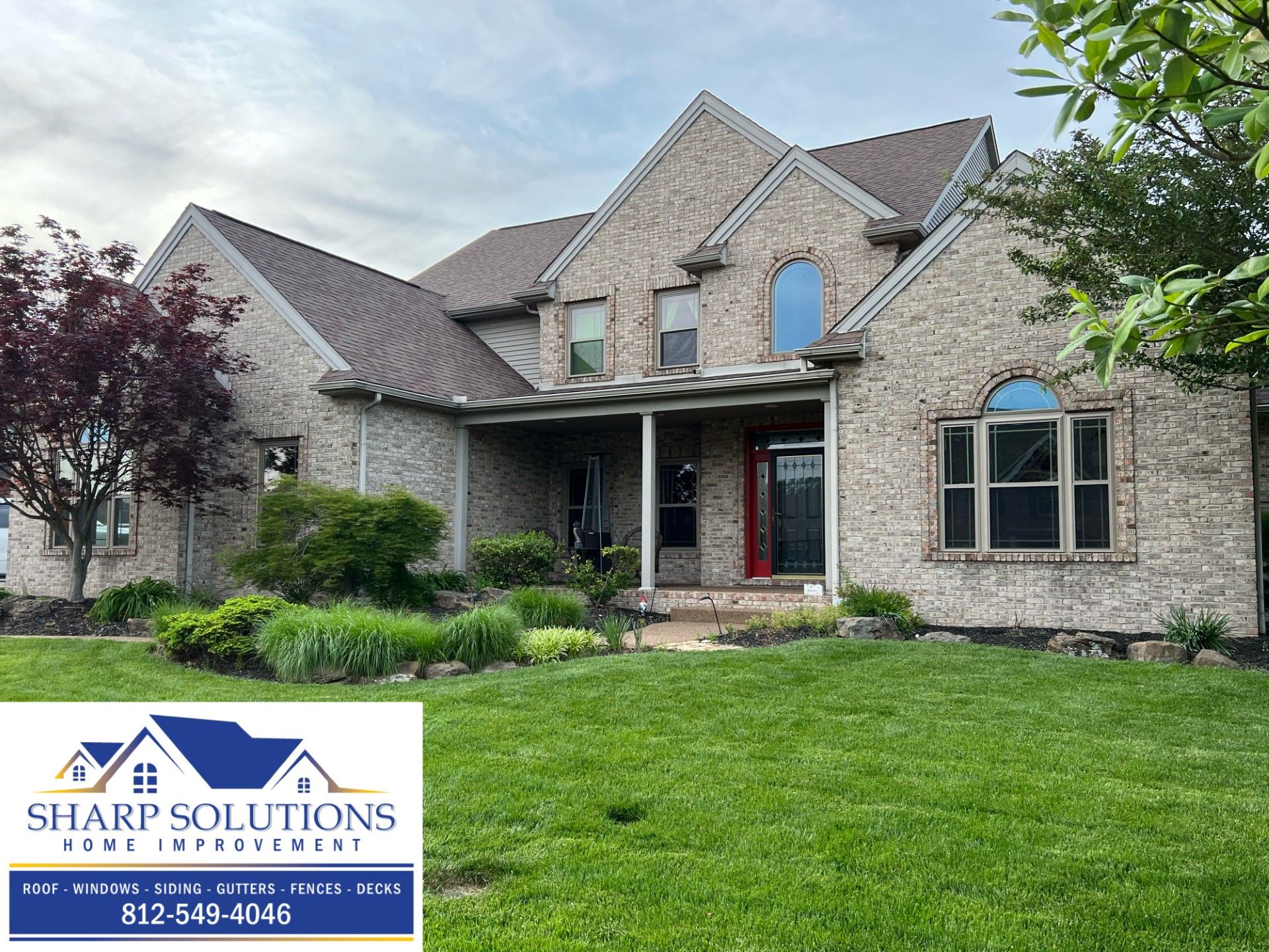 Stone suburban house with landscaped front yard and Sharp Solutions roofing logo in the corner