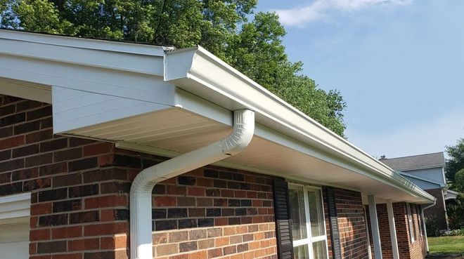 Brick house eave with white gutter and downspout under a clear blue sky.