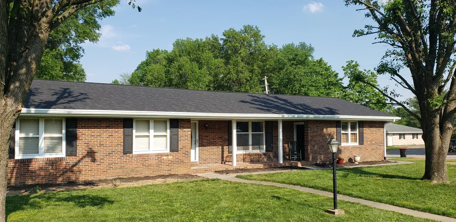 Single-story brick house with a dark roof, white shutters, and a front porch in a leafy yard.