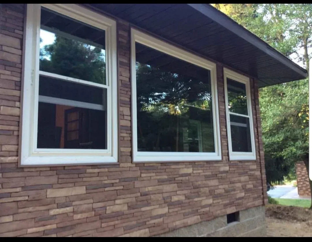 Brick house exterior with three large windows and white trim beside a wooded yard