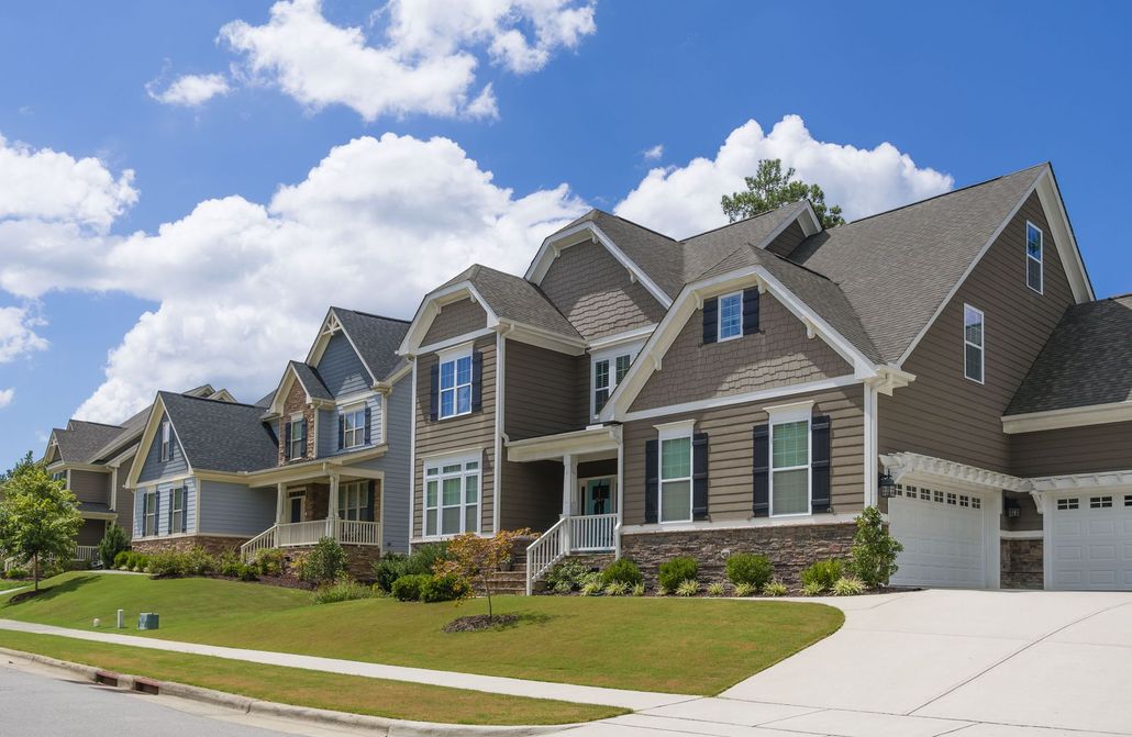 Large suburban brick houses with gabled roofs, a driveway, and a blue sky with clouds