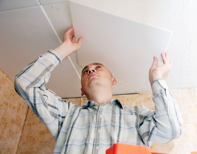 Man inspecting a ceiling tile with both hands raised in a tiled room