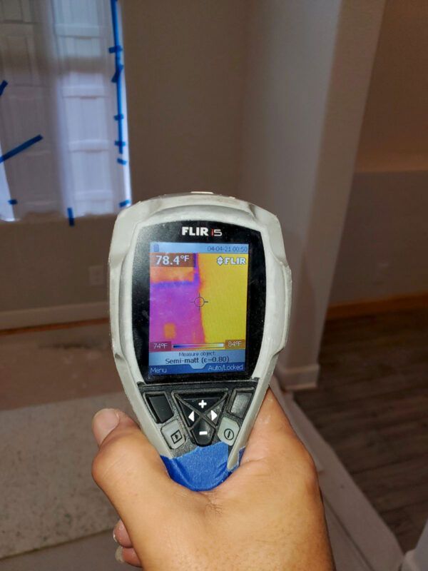 A hand holds a FLIR thermal imaging camera pointed at a wall, showing a temperature gradient between cool and warm areas.