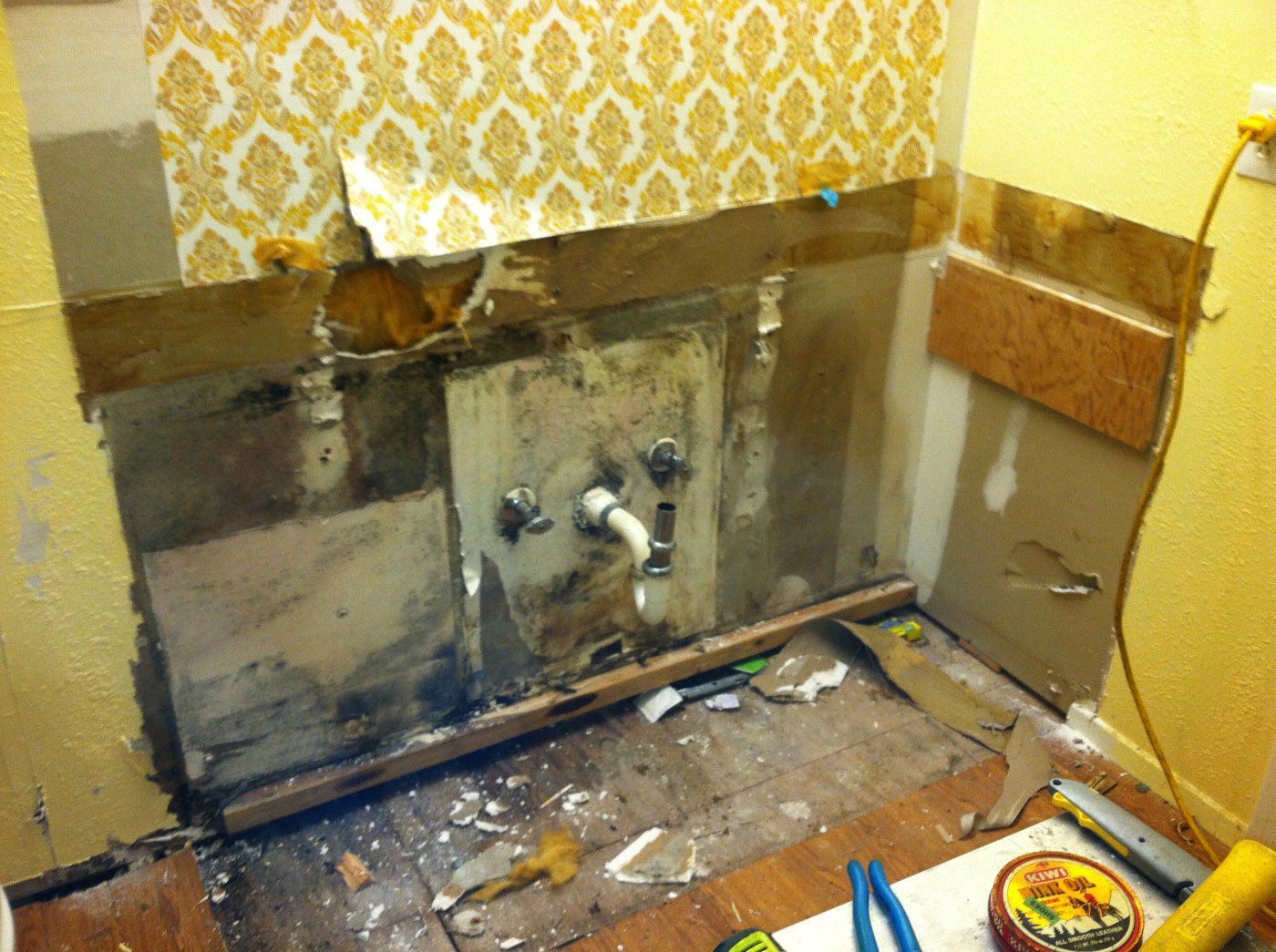 A bathroom sink area under renovation, featuring exposed walls, moldy drywall, visible plumbing, and yellow patterned paper.