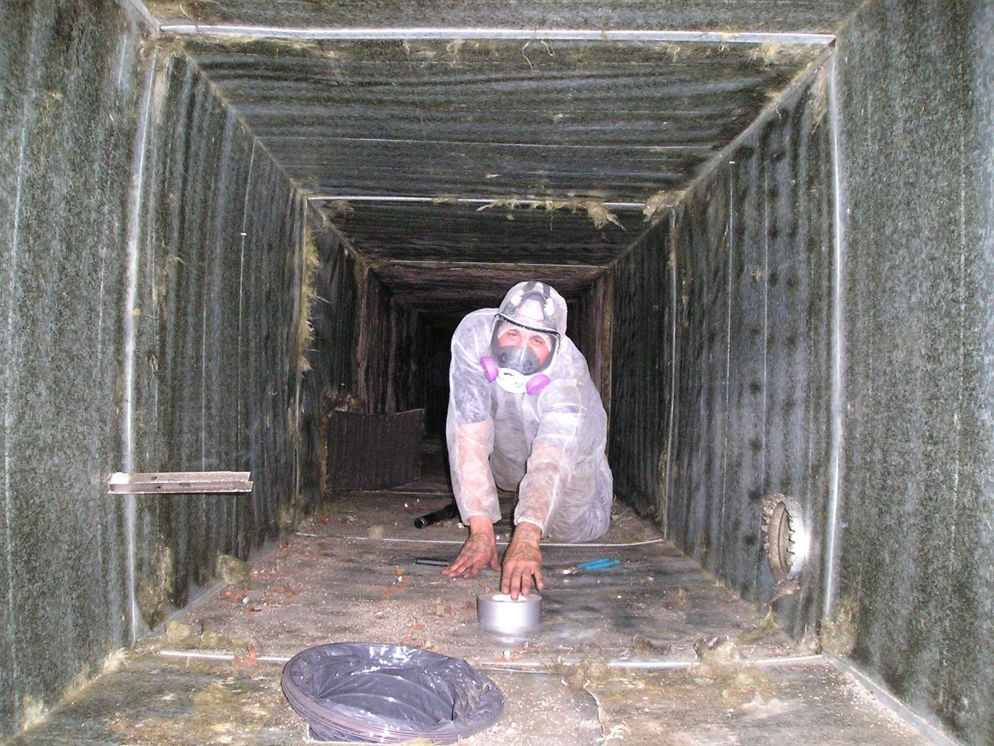 A technician in a white protective suit and respirator crawls through a dark, dusty rectangular air duct.