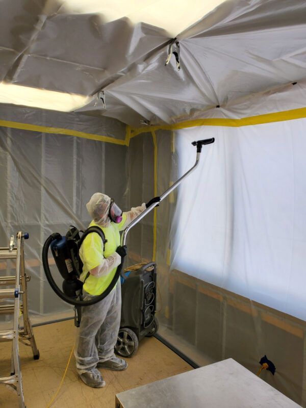 A person in protective gear and a respirator uses a backpack vacuum to clean the plastic-enclosed walls of a room.