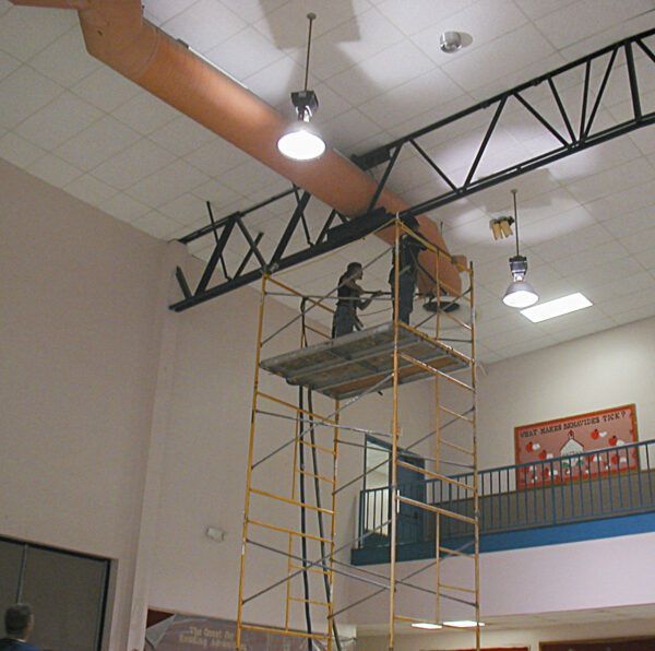 Two workers stand on high yellow scaffolding to perform maintenance on a large ceiling duct in a school hall.