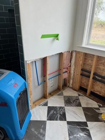 A room under repair showing exposed wooden studs, red and blue plumbing pipes, insulation, and a checkered marble floor.