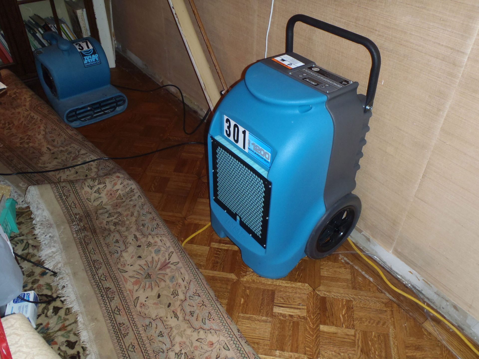 A blue professional-grade dehumidifier and a floor fan placed on a wooden floor in a room undergoing water damage repair.